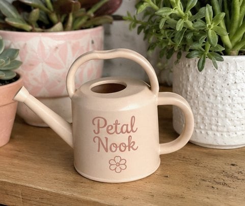 Small pastel mini indoor watering can with a generic boutique logo beside a potted plant